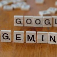 Scrabble tiles spelling "GEMINI" in front of blurred "GOOGLE".